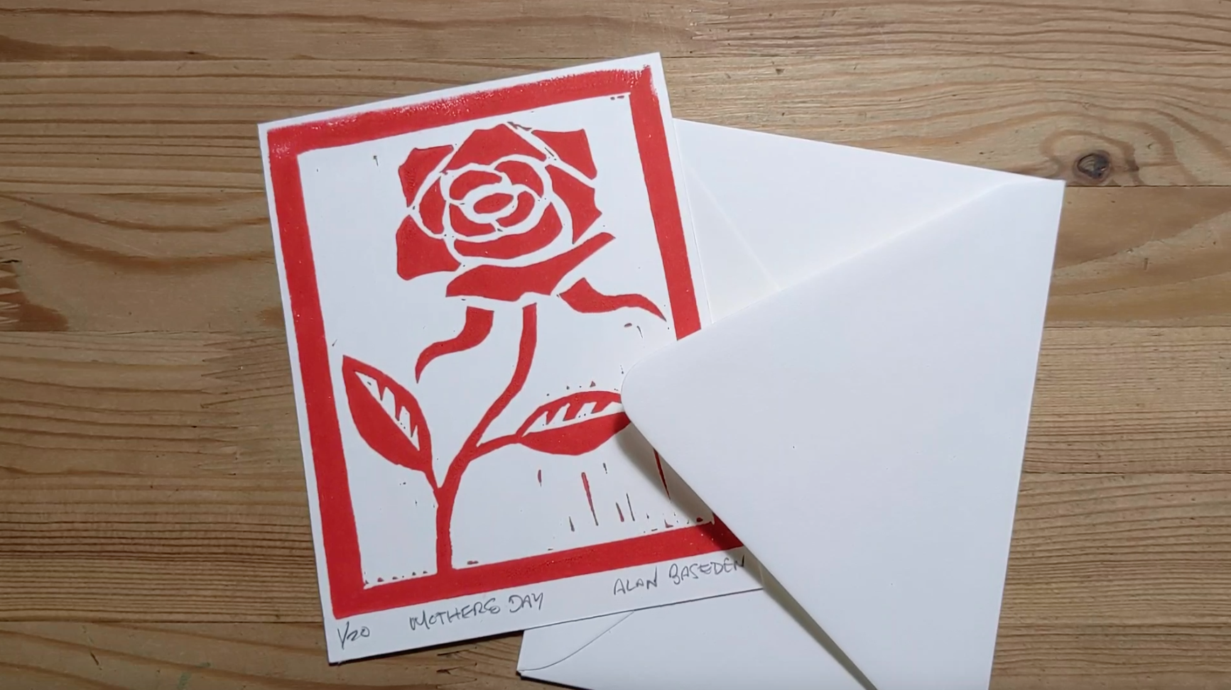 A greeting card with a red rose linocut print, and an envelope, on a wood table